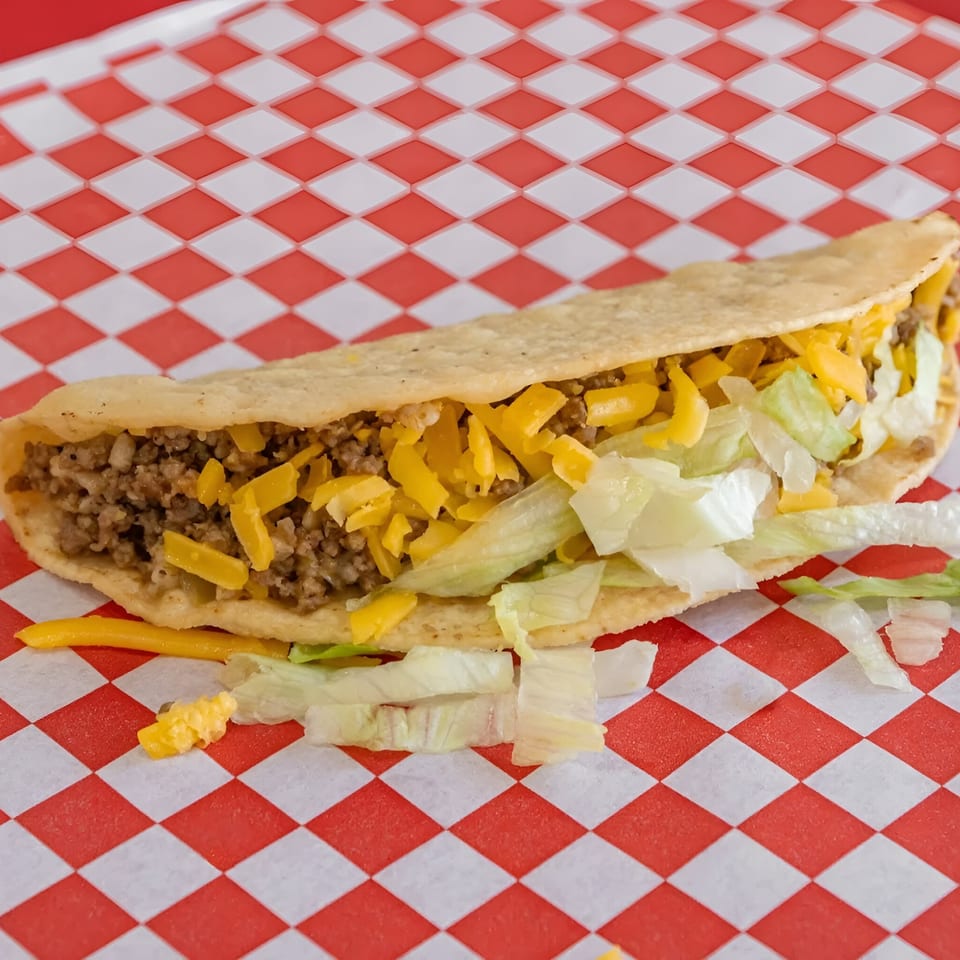 A crispy taco filled with ground beef, shredded cheddar cheese, and lettuce on red and white checkered paper.